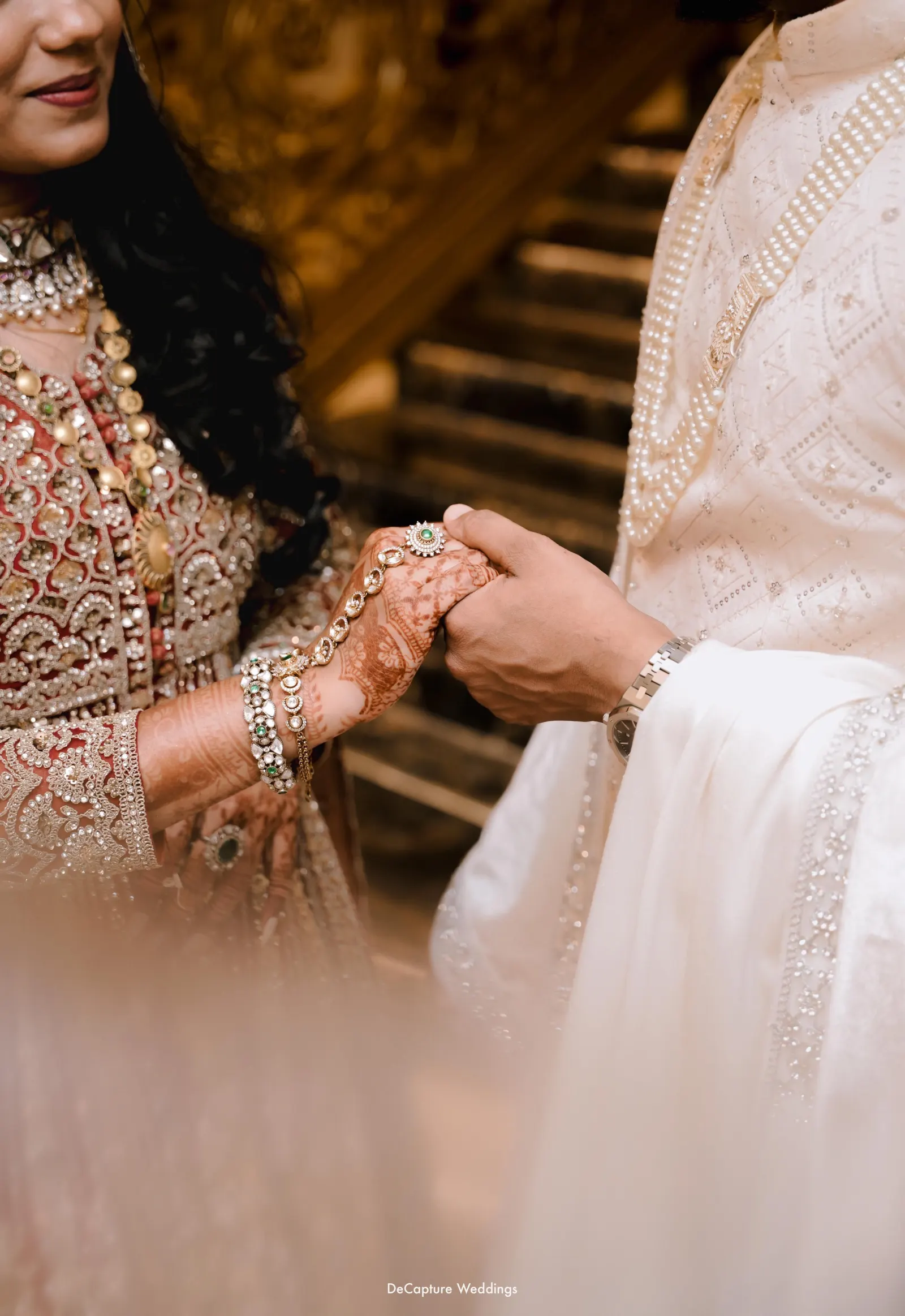 Close detail of the couple clasping hands with bridal henna, pearl strands, and intricate embroidery.