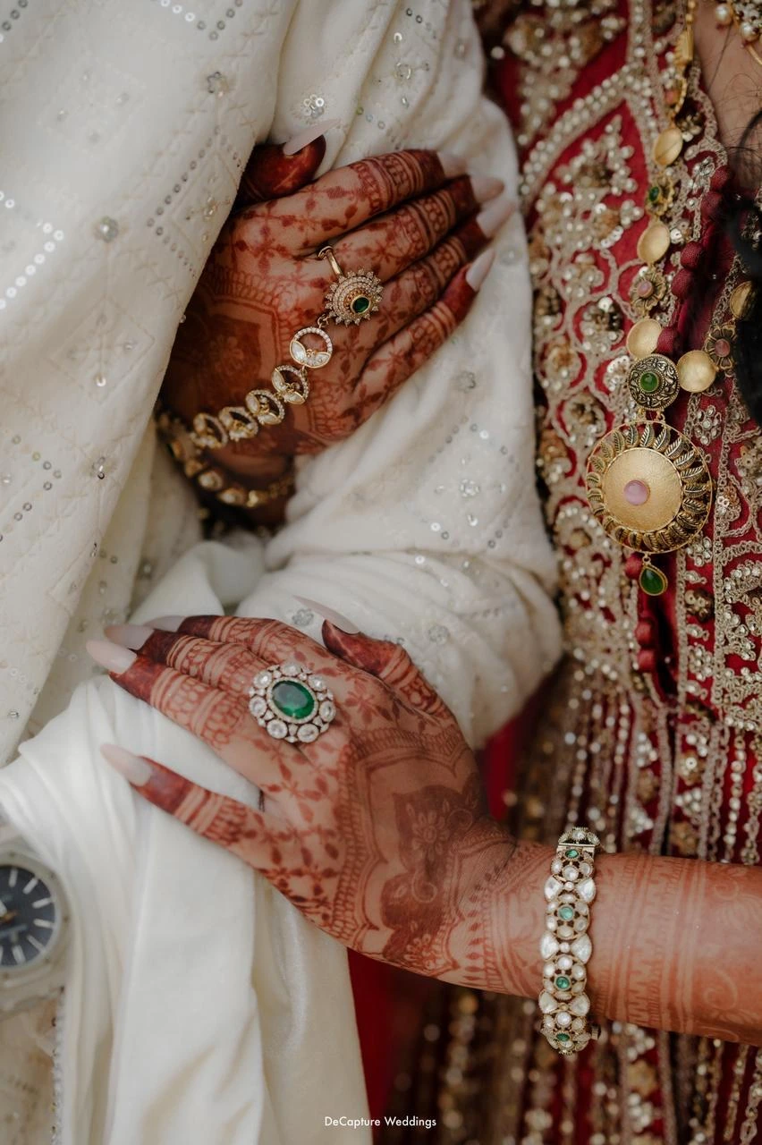 Intimate close-up of Mufeeda tenderly resting both hands on Ashhar's arm, adorned with intricate bridal henna, an emerald statement ring, and heirloom polki jewellery.